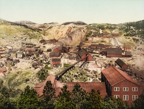 Aerial view of a historic mining town with buildings, machinery, and surrounding hills, showcasing a vibrant landscape.