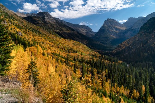 A scenic valley filled with vibrant autumn foliage, surrounded by mountains under a partly cloudy sky.