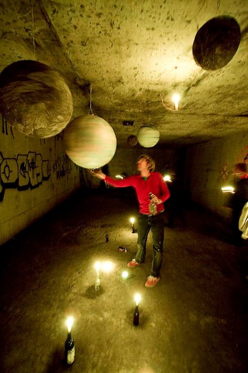 A person in a red shirt reaches for hanging planets in a dimly lit underground space, surrounded by candles.