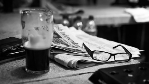 A glass of dark beer beside a stack of newspapers and a pair of glasses on a table.