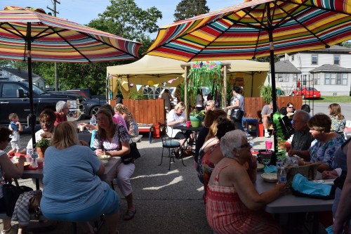 A lively outdoor gathering with people sitting at tables under colorful umbrellas, enjoying food and conversation.