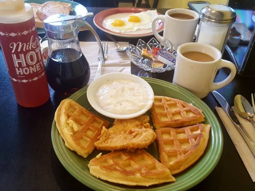A plate of waffles with fried chicken, a bowl of white gravy, coffee, and syrup on a table with breakfast items in the background.