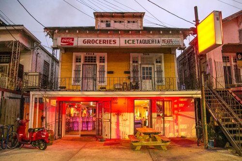 A colorful two-story building with a grocery store and taqueria, illuminated by warm lights at dusk.