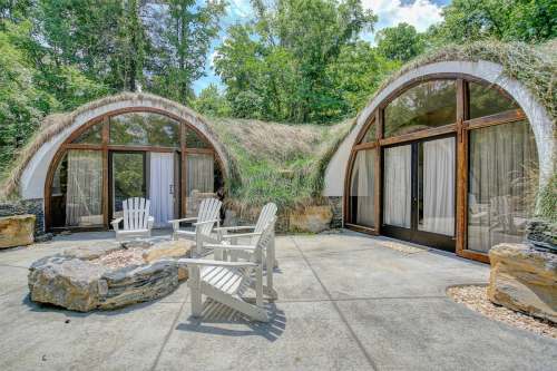 Two unique, curved buildings with grass roofs surrounded by trees, featuring a stone patio and wooden chairs.
