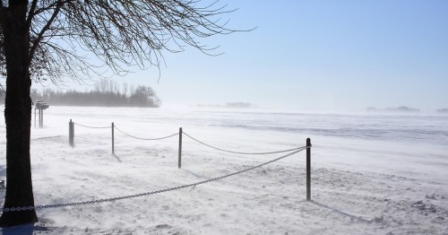 A snowy landscape with a chain-link fence, bare trees, and a misty horizon under a clear blue sky.