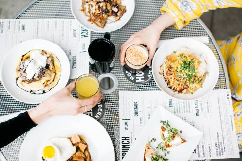 A table set with various brunch dishes and drinks, including pancakes, eggs, and cocktails, with hands reaching for food.