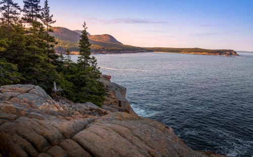 A scenic view of rocky coastline, trees, and calm waters under a colorful sky at sunset.