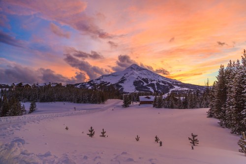 A snowy landscape at sunset, featuring a mountain in the background and colorful clouds in the sky.