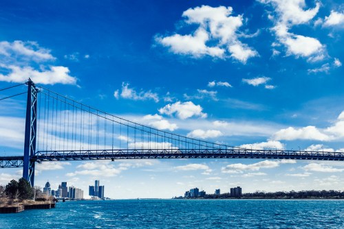 A blue sky with clouds above a large suspension bridge spanning a river, with a city skyline in the background.