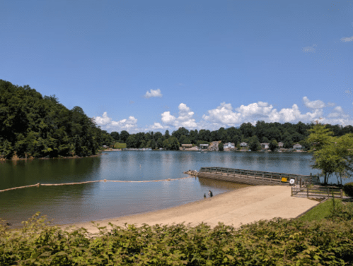 A serene lake scene with a sandy beach, green trees, and a clear blue sky dotted with clouds.