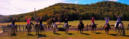 A group of eight people on horseback, raising their arms, with a scenic green hillside in the background.