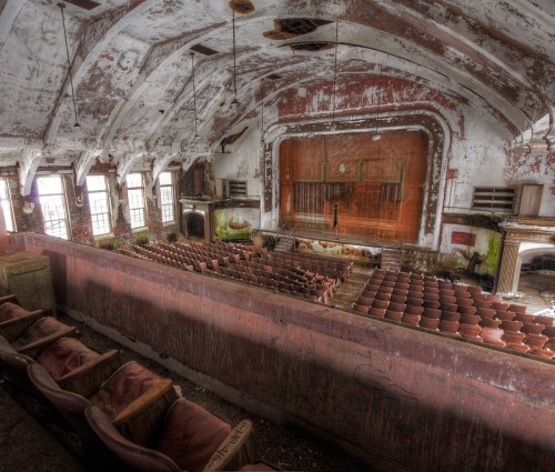 Abandoned theater interior with peeling paint, empty seats, and a large stage in disrepair.