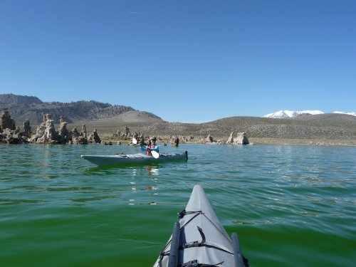 A view from a kayak on a calm lake, with another kayaker in the distance and mountains in the background.
