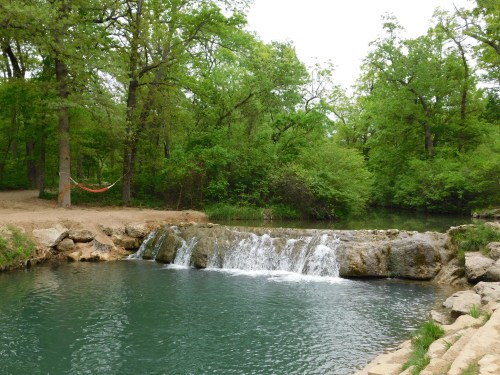 A serene scene of a small waterfall flowing into a calm, greenish-blue pond, surrounded by lush trees and a hammock.