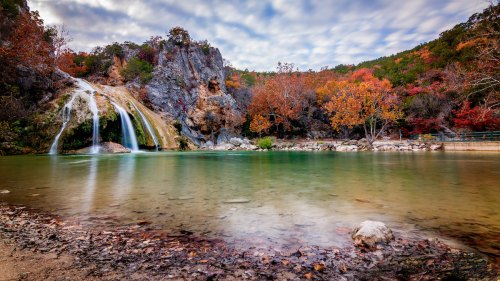 A serene waterfall cascades into a tranquil pool, surrounded by autumn foliage and rocky cliffs under a cloudy sky.