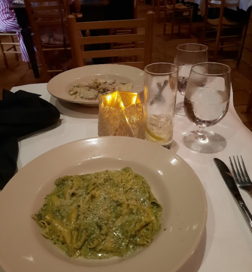 A plate of pesto pasta sits on a table with a candle, water glasses, and another dish in the background.