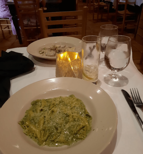 A plate of pesto pasta sits on a table with a candle, water glasses, and another dish in the background.