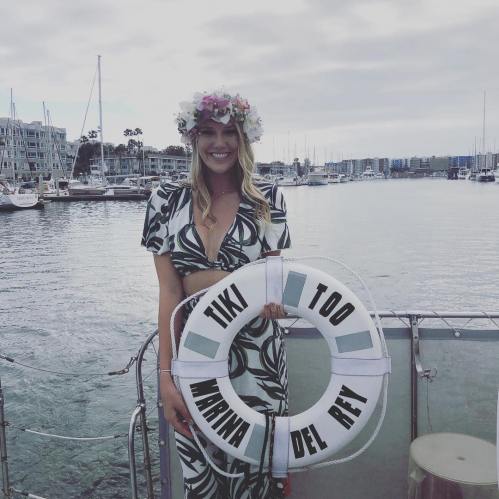 A smiling woman in a floral crown holds a life ring that reads "Tiki Too Marina Del Rey" by a marina.