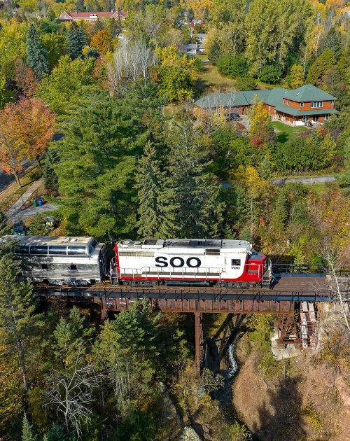 A train crosses a wooden bridge surrounded by colorful autumn trees and houses in the background.