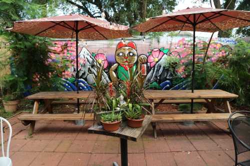 A vibrant outdoor seating area with two umbrellas, a mural of a character, and potted plants on a table.