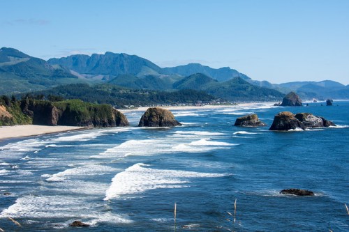 Scenic coastal view with waves, rocky formations, and mountains in the background under a clear blue sky.