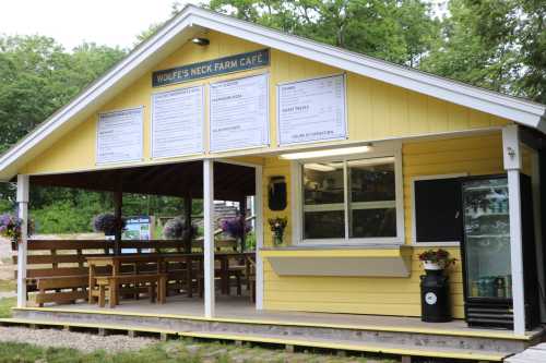 A yellow café building with a menu board, outdoor seating, and a fridge, surrounded by greenery.