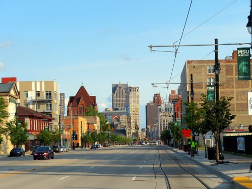 A city street lined with buildings, trees, and a clear blue sky, leading towards a skyline of tall buildings.