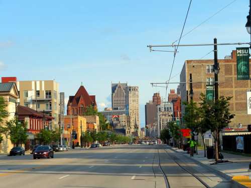 A city street lined with buildings, trees, and a clear blue sky, leading towards a skyline of tall buildings.