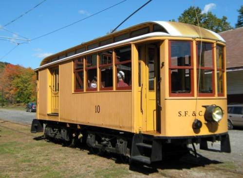 A vintage yellow streetcar with large windows, parked on a gravel path surrounded by trees and blue sky.