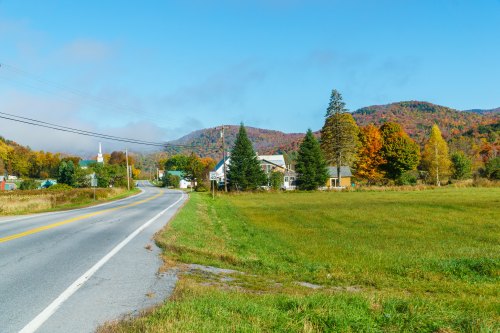 A scenic rural road lined with trees and colorful autumn foliage, leading to a small town with mountains in the background.