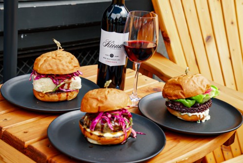 Three gourmet burgers on black plates, accompanied by a glass of red wine, set on a wooden table.