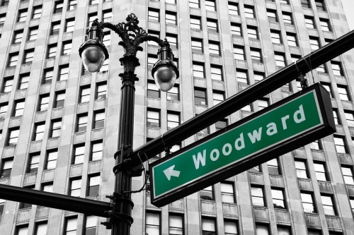 Green street sign for "Woodward" with an arrow, against a black and white backdrop of a tall building.