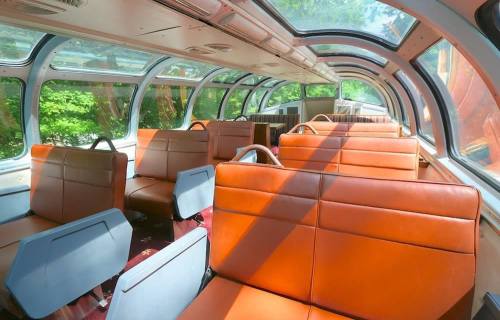 Interior of a train car with large windows, featuring orange leather seats and a bright, sunny atmosphere.