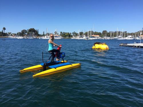 A person in a blue tank top rides a yellow pedal boat on a calm waterway with boats in the background.