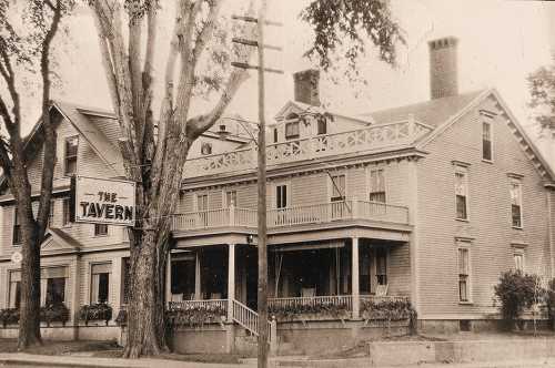 Historic building with a sign reading "TAVERN," featuring a porch and large trees in front. Black and white photo.