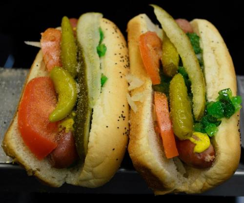 Two hot dogs in buns, topped with pickles, tomatoes, mustard, and green relish.