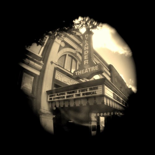 A vintage theater marquee displaying "Flander Theatre" with a musical title, framed by trees and a cloudy sky.