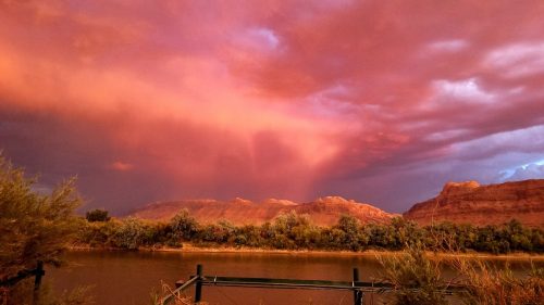 A vibrant sunset casts pink and orange hues over a river, with mountains and lush greenery in the foreground.