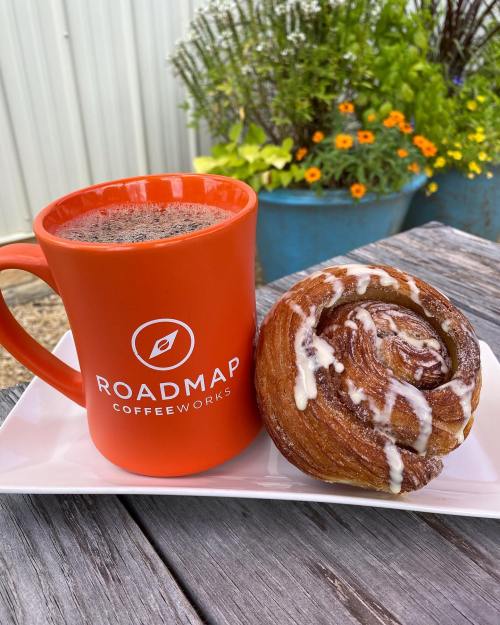 A bright orange coffee mug beside a cinnamon roll on a white plate, with flowers in the background.