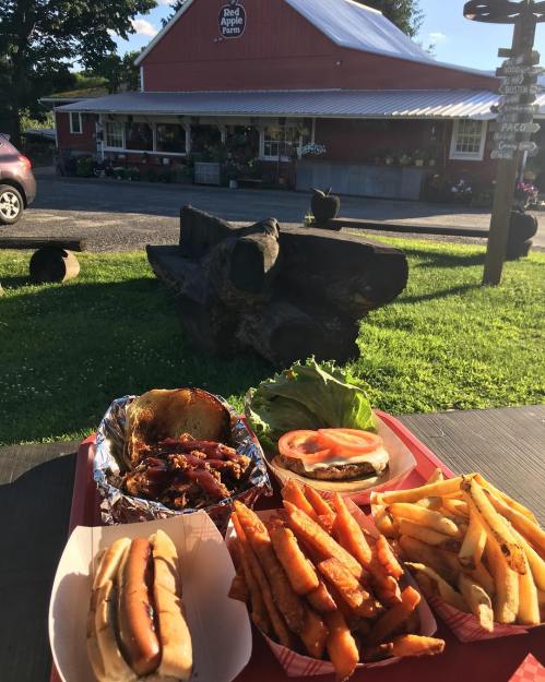 A tray of hot dogs, a burger with lettuce and tomato, and sweet potato fries in front of a red barn restaurant.