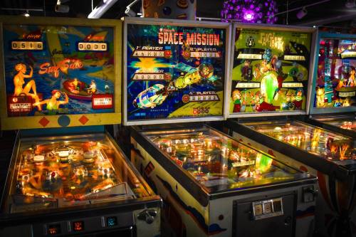 A vibrant arcade scene featuring three vintage pinball machines with colorful graphics and illuminated playfields.