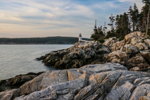 A rocky coastline with a lighthouse in the distance, surrounded by trees and calm waters under a cloudy sky.