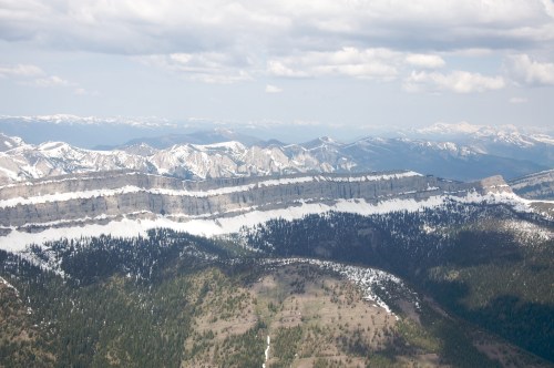 Aerial view of snow-capped mountains with a rugged landscape and scattered greenery under a cloudy sky.