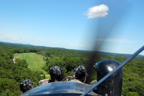 View from a vintage aircraft cockpit, showcasing a green landscape and blue sky with a few clouds.