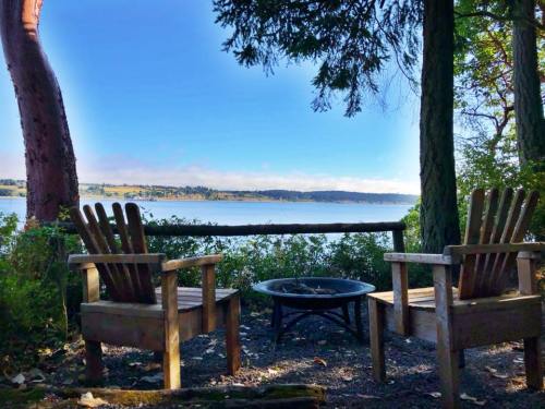 Two wooden chairs facing a fire pit, overlooking a serene lake surrounded by trees and a clear blue sky.