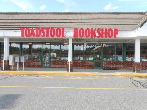 Exterior of Toadstool Bookshop, featuring a red sign and large windows displaying books, set in a shopping plaza.