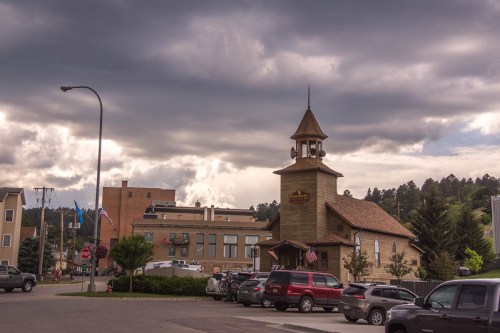 A quaint building with a clock tower, surrounded by cars and trees, under a cloudy sky in a small town.