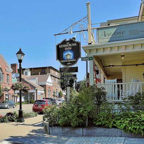 A street view featuring the Kennebunk Inn sign, nearby buildings, and a lamppost with flowers in a charming town setting.