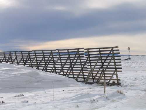 A wooden fence stretches across a snowy landscape under a cloudy sky, with a windmill visible in the distance.