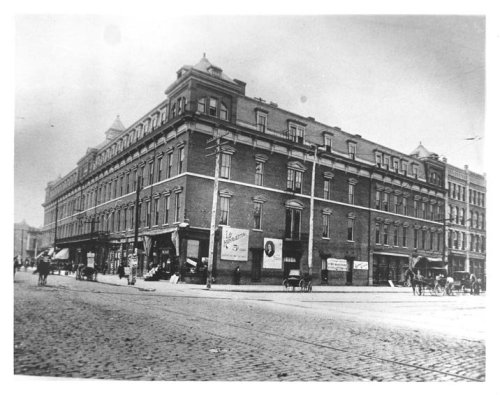 Historic black-and-white photo of a large brick building on a street corner, with horse-drawn carriages nearby.